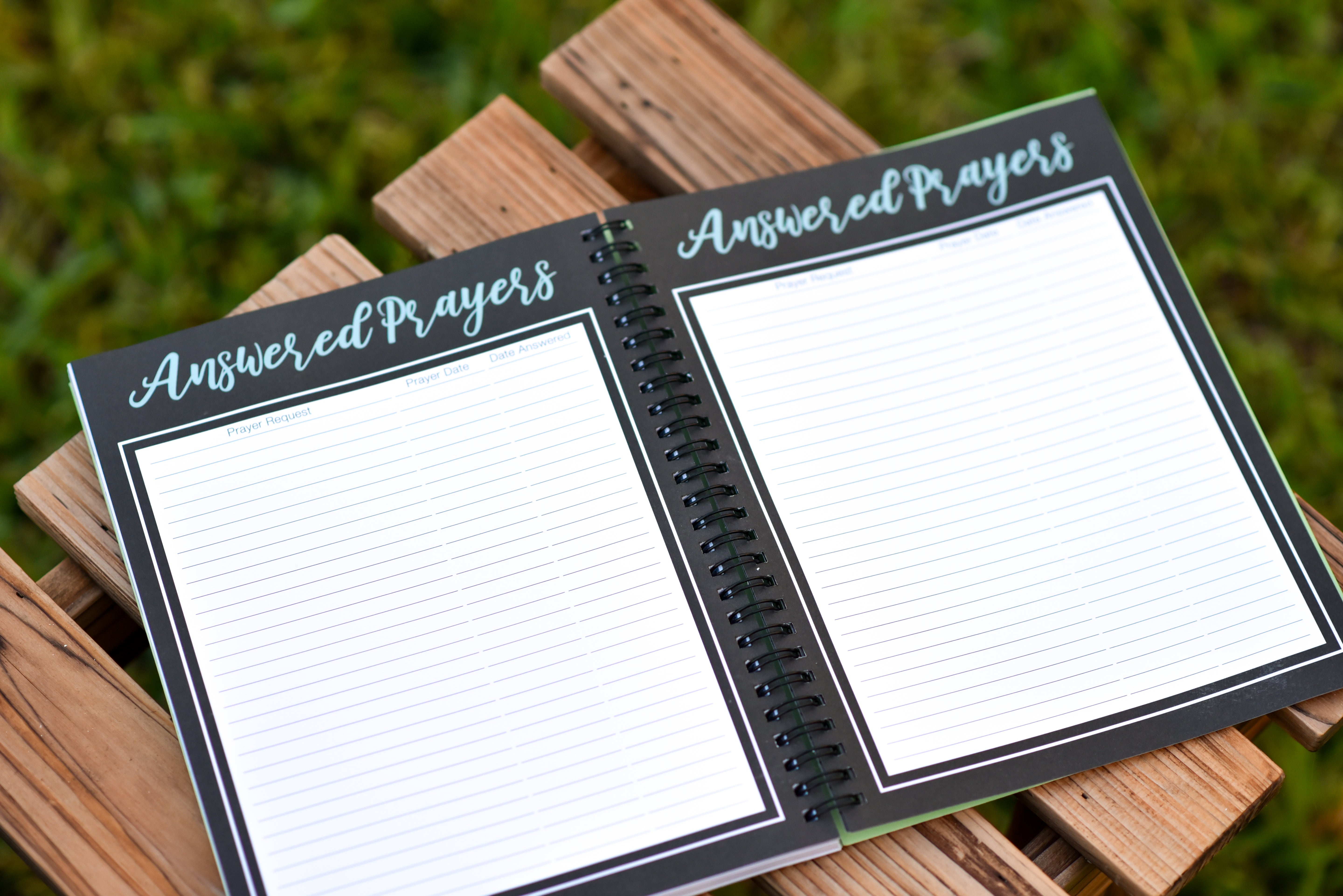 Open spiral-bound notebook titled 'Answered Prayers' on a wooden surface with grass in the background.