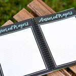Open spiral-bound notebook titled 'Answered Prayers' on a wooden surface with grass in the background.
