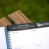 Notebook with a decorative border featuring a Bible verse scripture and illustrations on a wooden surface.