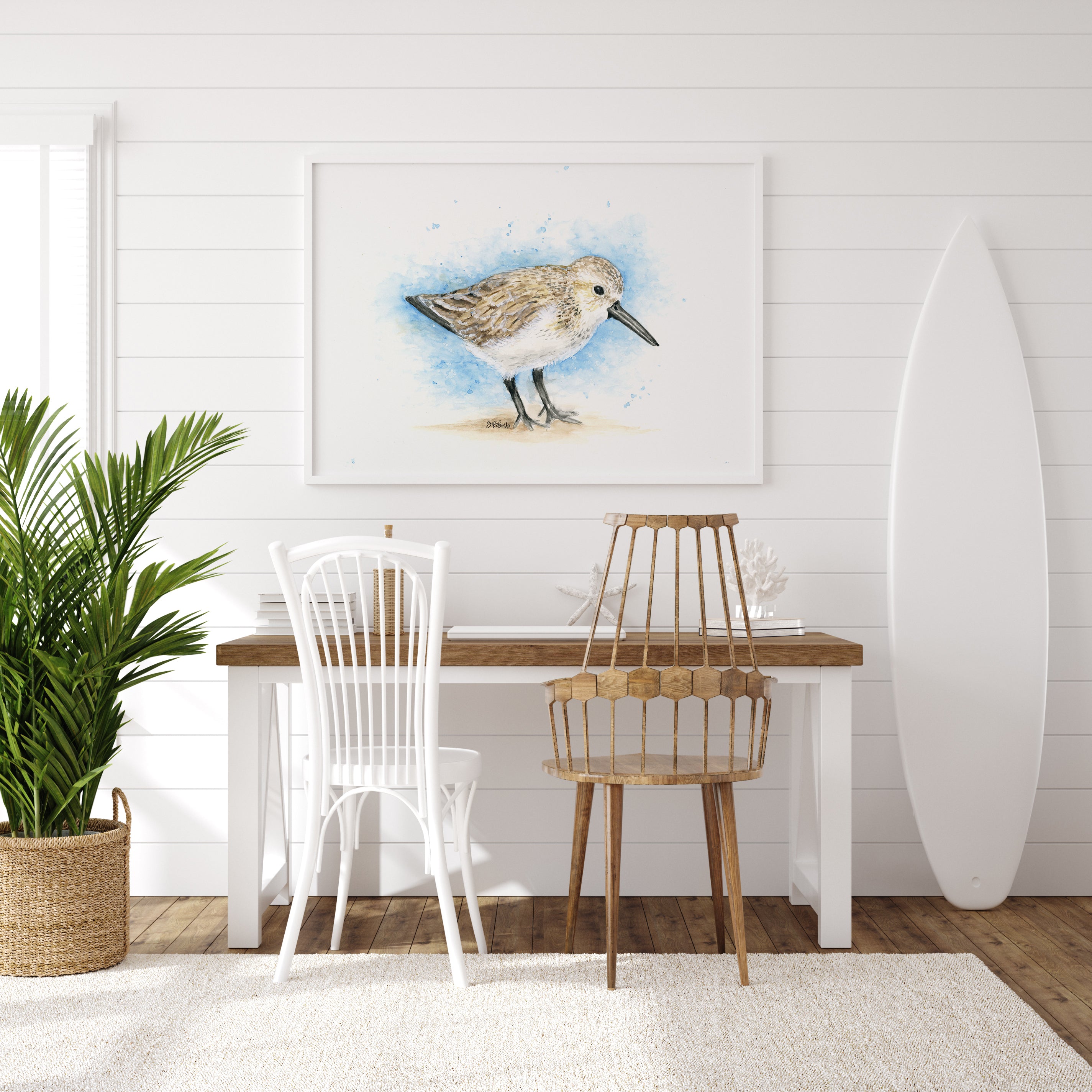 Dining area with a table, chairs, and a surfboard against a white wall with a sandpiper bird painting.