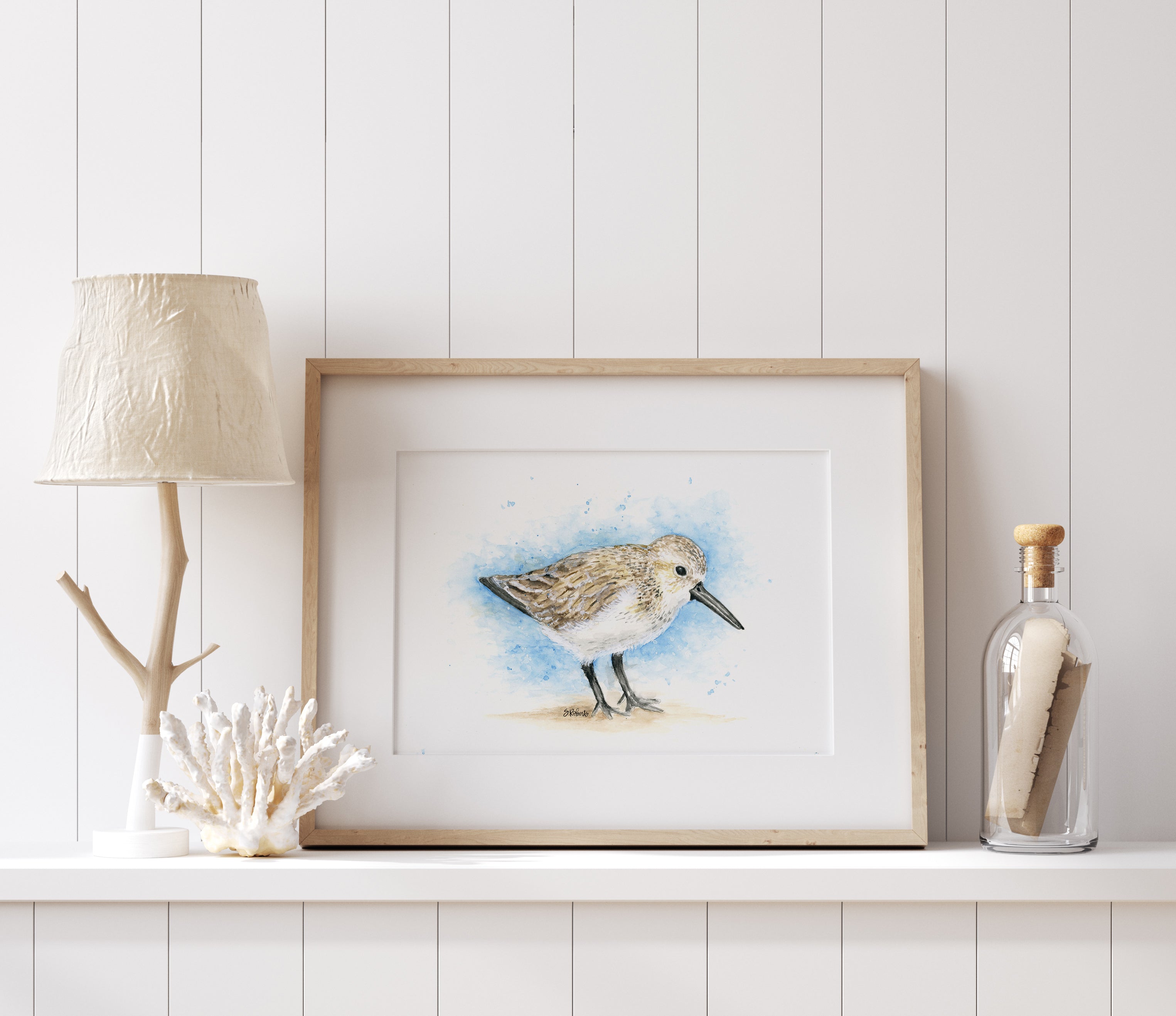 Framed artwork of a bird on a shelf with decorative items against a white wall.